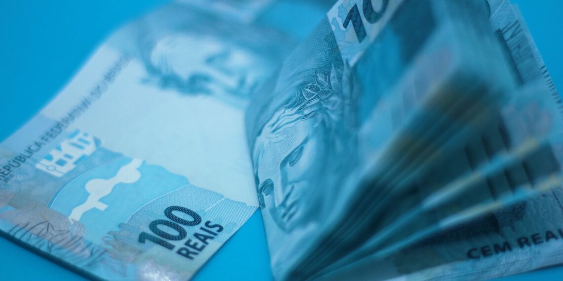Pilha de cédulas de 100 reais sobre superfície azul, ilustrando a inflação medida pelo IPCA-15